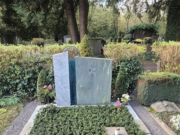 Grabstätte mit einem polierten grauen Grabstein auf einem Friedhof, umgeben von Kies, Blumen und Bäumen.