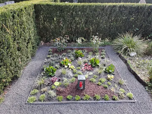 Grab mit bunten Blumen und Grünpflanzen, eingefasst von Kies und Hecken.