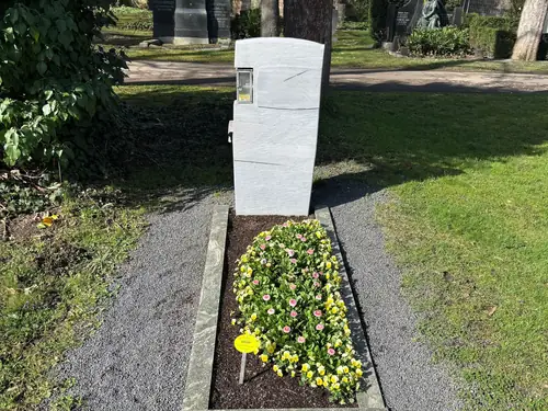 Grabstein mit weißer Gedenktafel und Blumenbeet auf einem Friedhof im Sonnenlicht