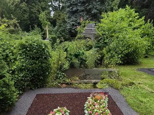 Blumenbeet als Gedenkstätte auf einem Friedhof, mit farbenfrohen, herzförmigen Blumen in einem dunklen Kiesrand.