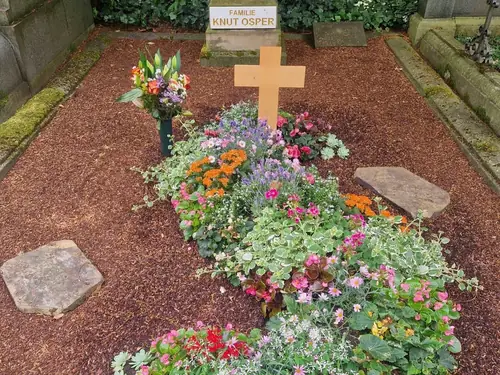 Ein mit Blumen bedecktes Grab mit einem Holzkreuz und einer Gedenktafel in einem Friedhof