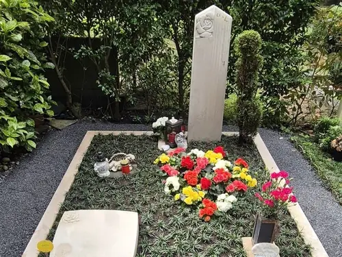 Kleiner Grabstein mit Blumen auf einem Friedhof, umgeben von Kies und Hecken.