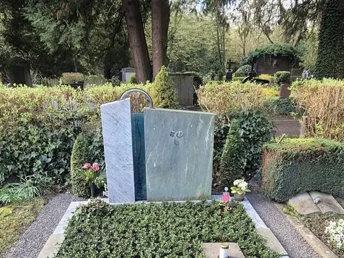 Grabstätte mit einem polierten grauen Grabstein auf einem Friedhof, umgeben von Kies, Blumen und Bäumen.